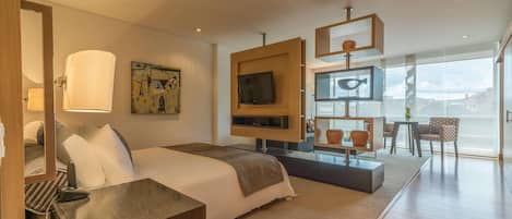 Une chambre d’hôtel moderne avec un grand lit, un téléviseur fixé sur un panneau en bois, un bureau avec une lampe et une vue sur la ville grâce à de grandes fenêtres.