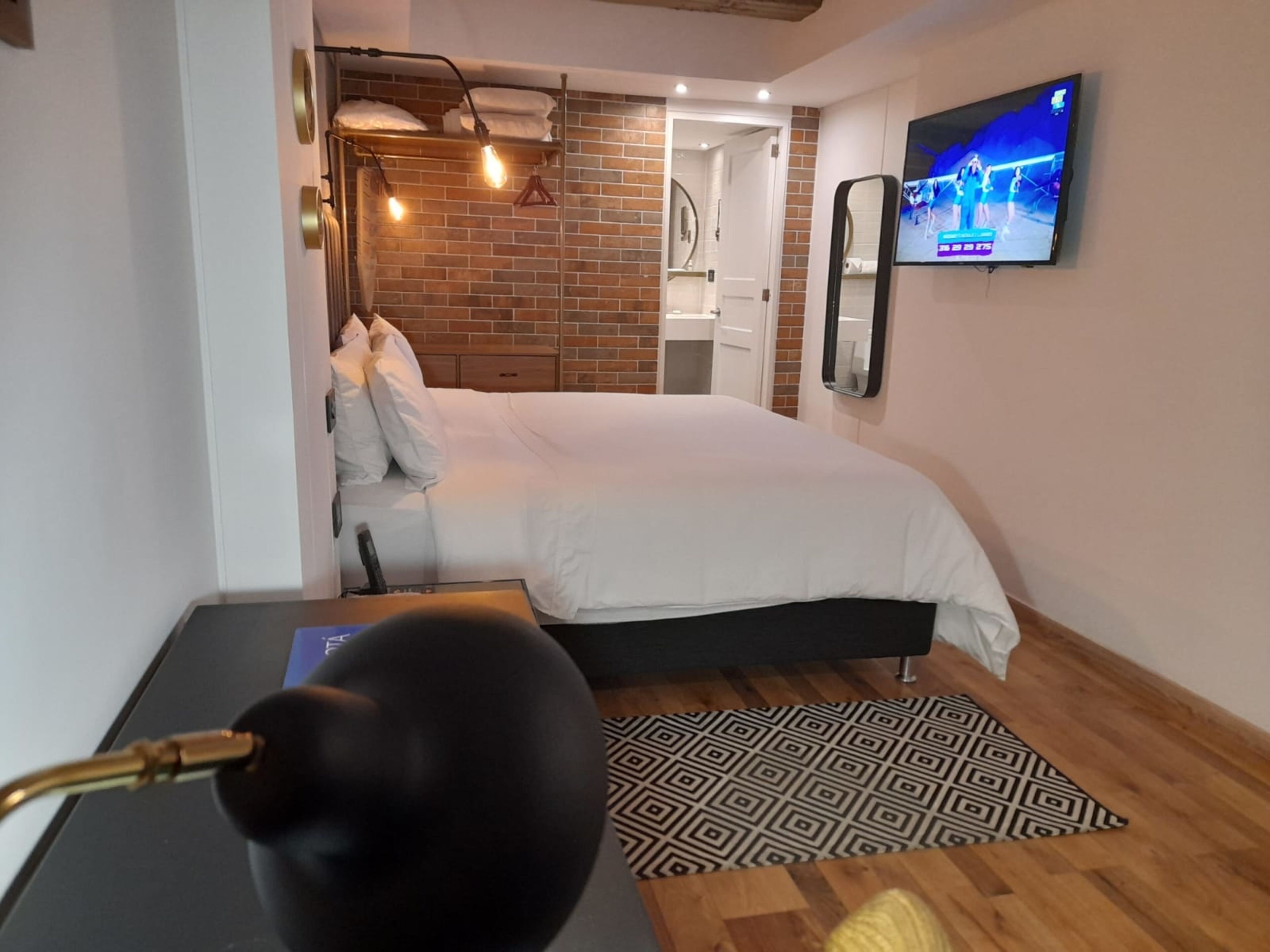 A modern hotel room with a large bed, a TV mounted on the wall, and a brick accent wall.