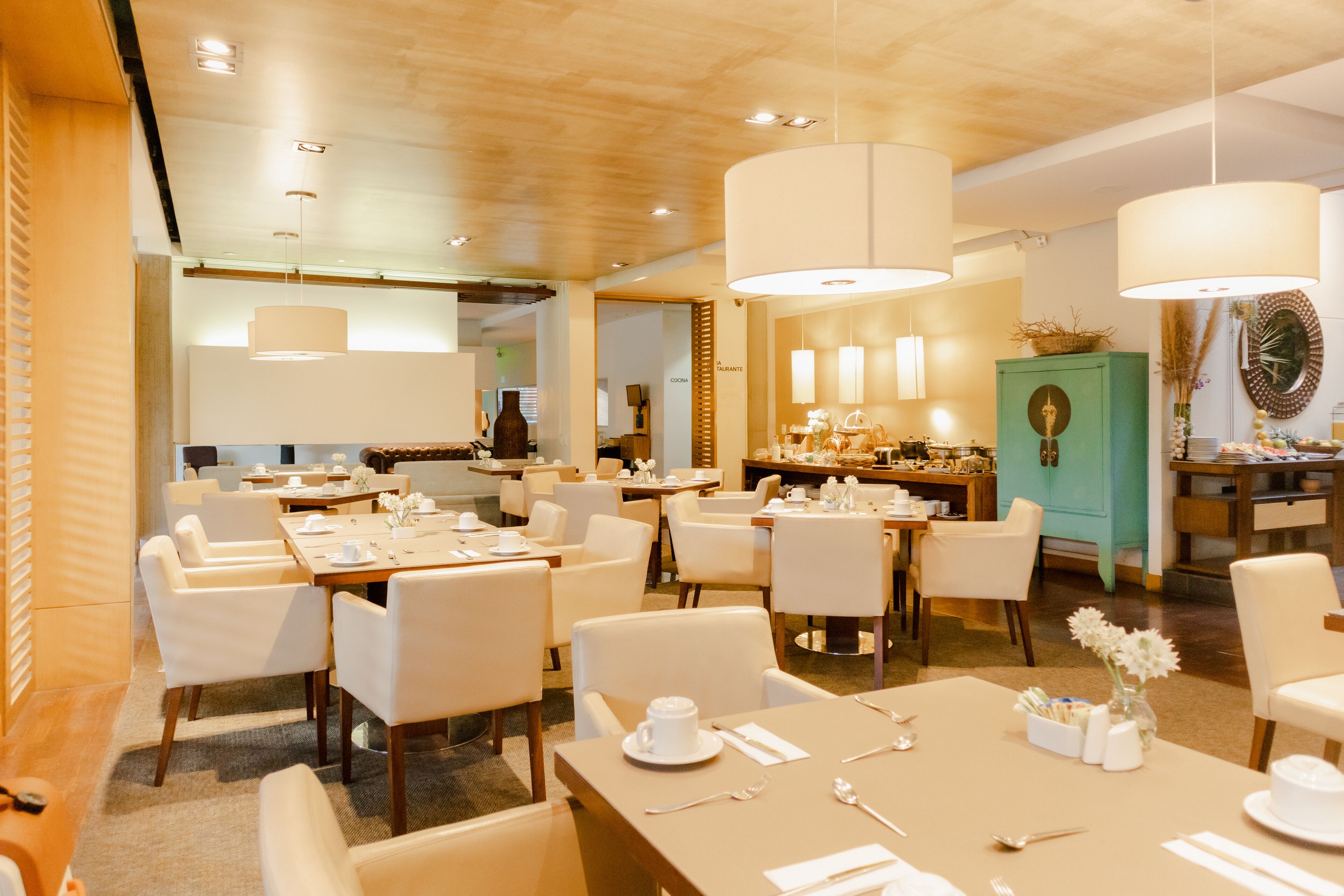 A modern hotel dining area with multiple tables and chairs, large pendant lights, and a green cabinet with decorative items.