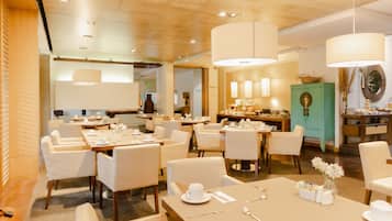 A modern hotel dining area with multiple tables and chairs, large pendant lights, and a green cabinet with decorative items.