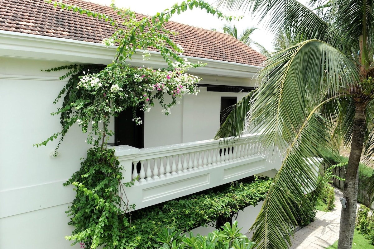 A white building with a red-tiled roof, a balcony with white balustrades, and lush greenery including a palm tree.