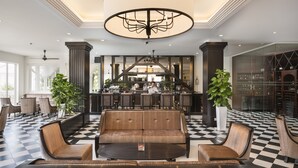 A lobby with a checkered floor, a central seating area with a sofa and chairs, a bar with a bartender, and a large chandelier.