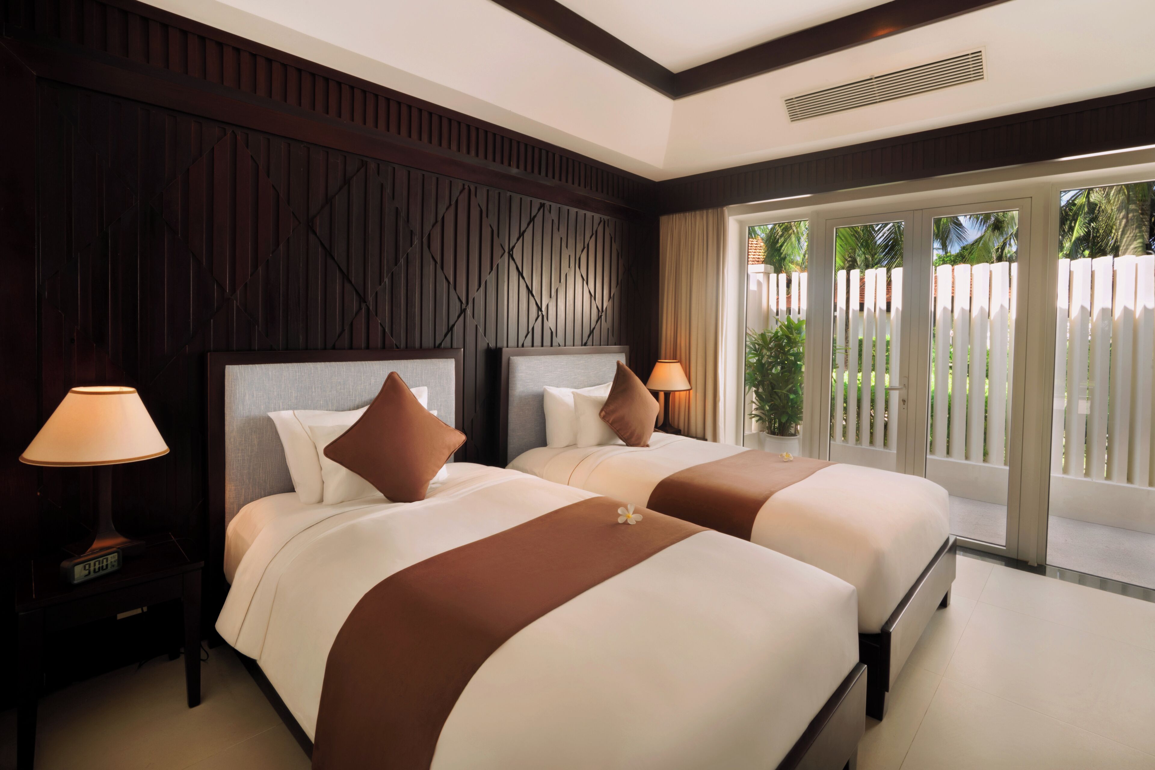 A hotel room with two beds, a nightstand with a lamp, and a view of a white fence and greenery outside.