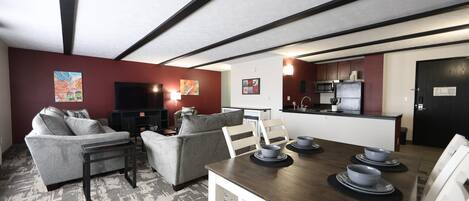 Ein modernes Wohnzimmer mit einer Couch, Sesseln, einem Couchtisch und einem Esstisch mit Schüsseln. Im Hintergrund ist eine Küche mit einer Mikrowelle und einem Kühlschrank erkennbar.
