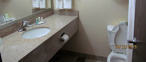 Un baño con lavamanos, espejo e inodoro. Hay toallas colgadas en la pared y un estante con más toallas.