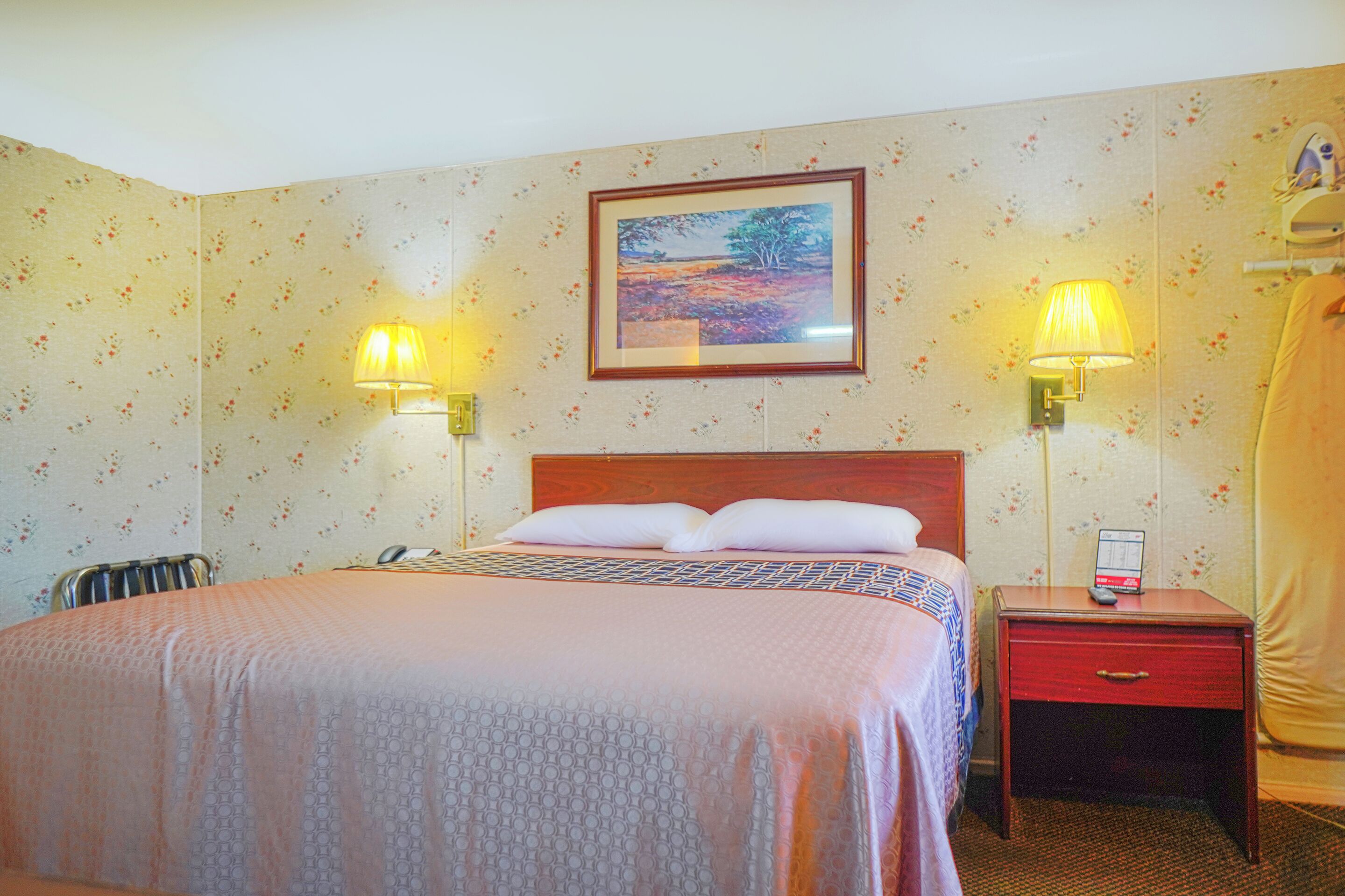 Habitación de hotel con cama, dos lámparas de noche, una mesita de noche con un teléfono y un cuadro en la pared.