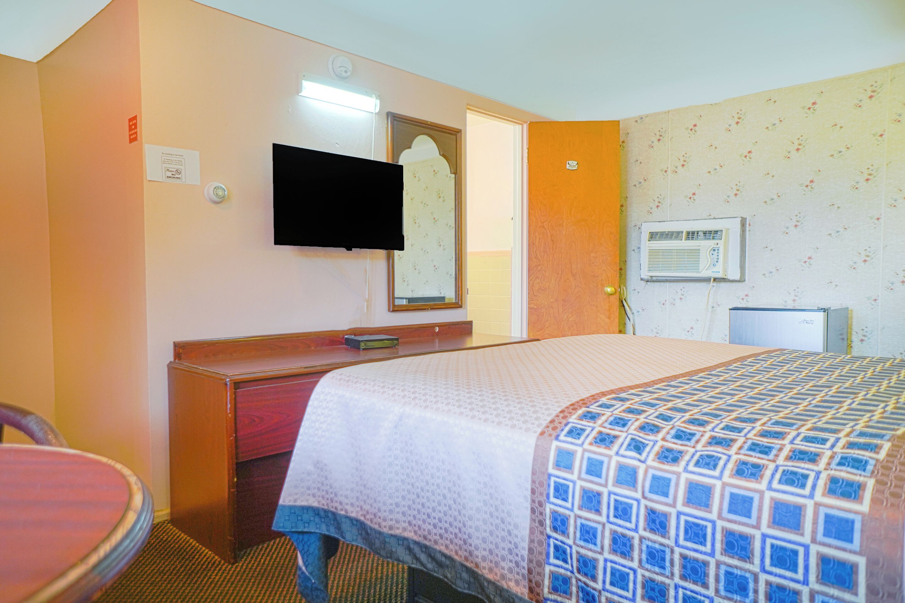 A hotel room with a bed, a desk, a chair, a television, and an air conditioning unit.