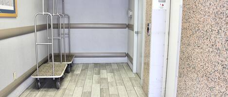 A hallway with a wooden floor, a door with a 'G' sign, and a wall-mounted fire alarm.