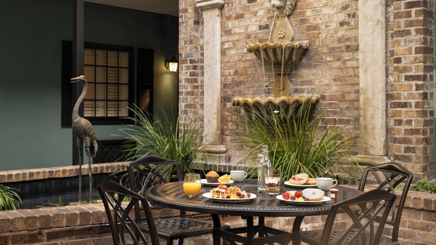 A dining table set with breakfast items, surrounded by chairs, in an outdoor patio area with brick walls and a decorative fountain.