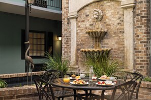 A dining table set with breakfast items, surrounded by chairs, in an outdoor patio area with brick walls and a decorative fountain. - Indigo Inn (Charleston)