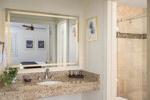 A bathroom with a granite countertop, a sink, a mirror, framed artwork, and a shower area.