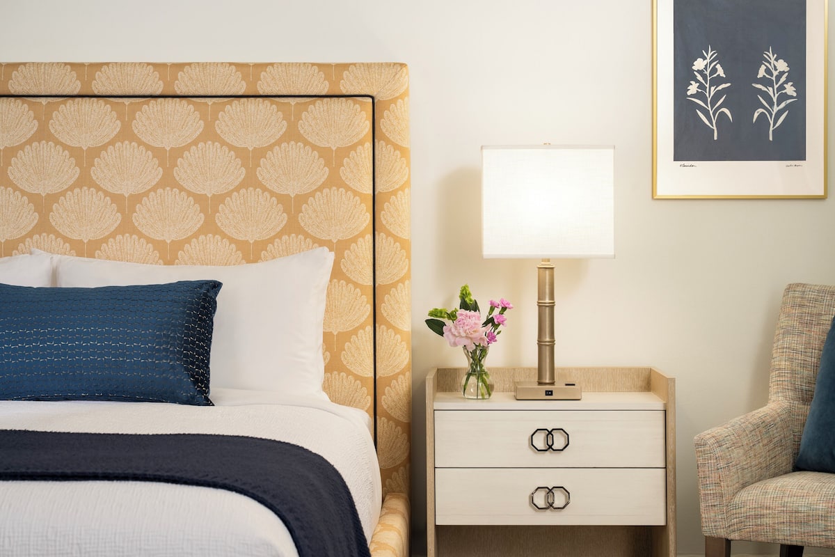 Habitación de hotel con un cabecero estampado, una mesita de noche con lámpara, una silla e impresión botánica enmarcada en la pared.