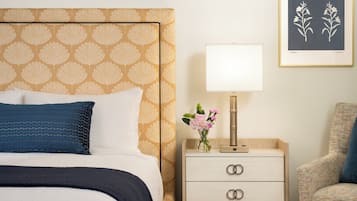 Quarto de hotel com cabeceira estampada, mesa de cabeceira com abajur, uma cadeira e uma impressão botânica emoldurada na parede.