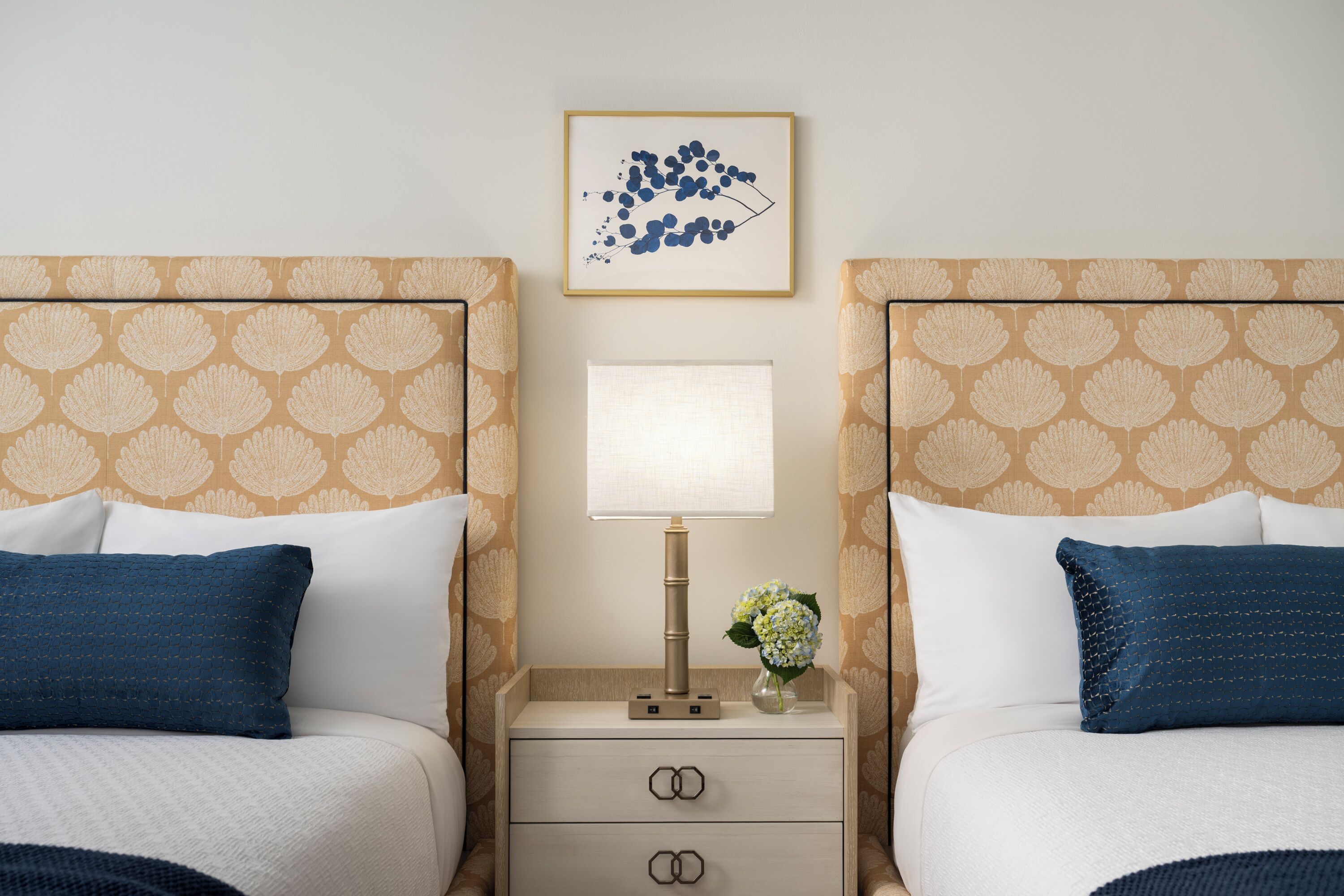 Une chambre d’hôtel avec deux lits, une table de chevet avec une lampe, un vase de fleurs et un tableau encadré au mur.
