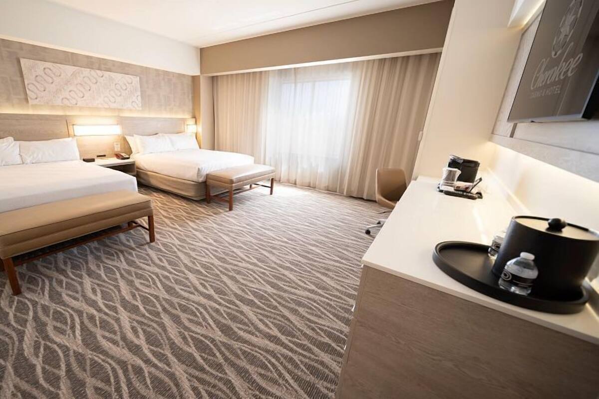A hotel room with two beds, a desk, a chair, and a large window with curtains.