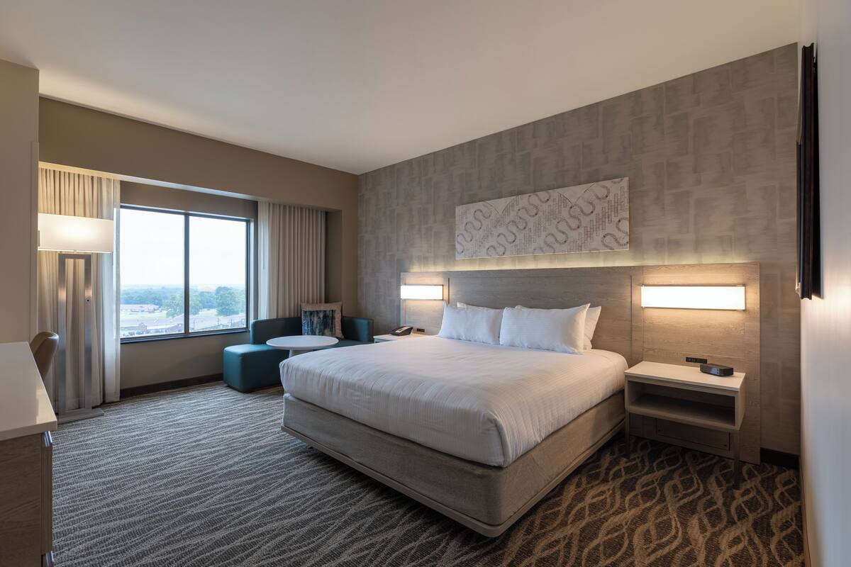 A hotel room with a large bed, a desk, a chair, a sofa, and a large window with curtains.
