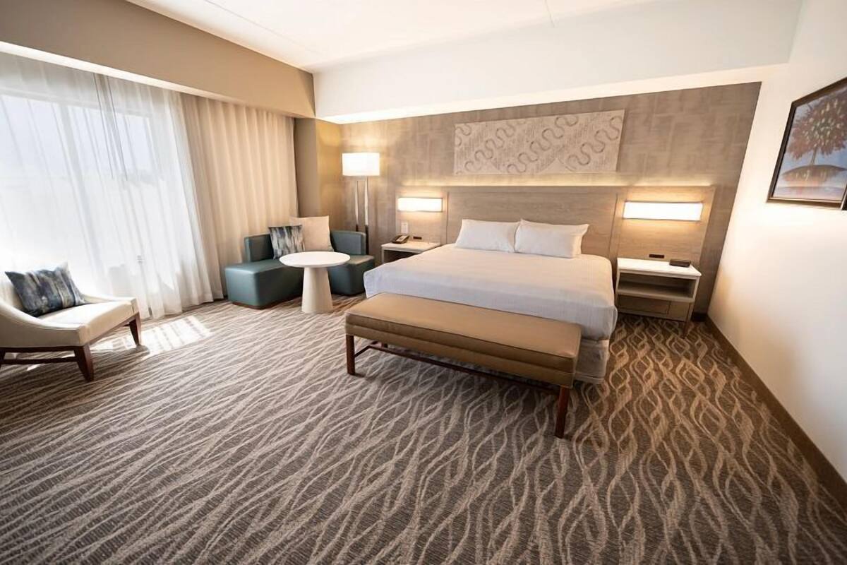 A hotel room with a large bed, a sofa, a chair, a small table, and a nightstand with a lamp.