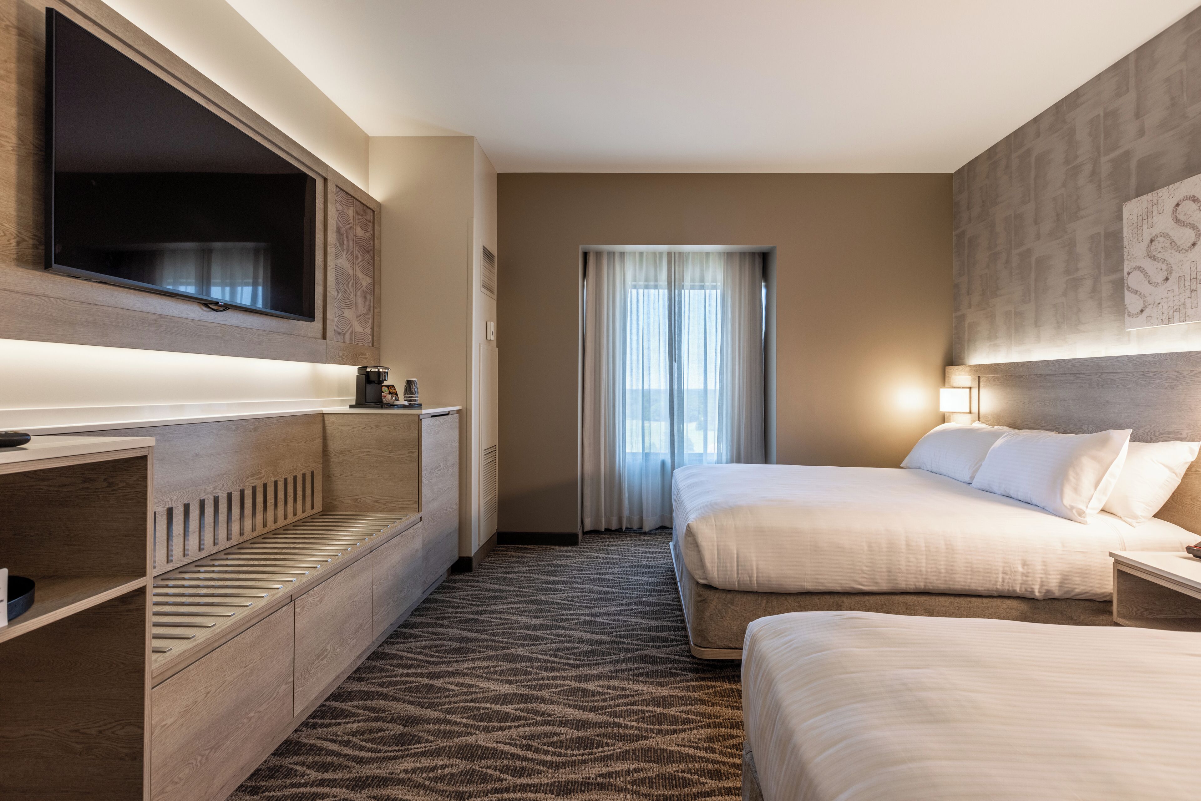 A hotel room with a large flat-screen TV mounted on the wall, a bed with white linens, a bedside table with a lamp, and a patterned carpet.