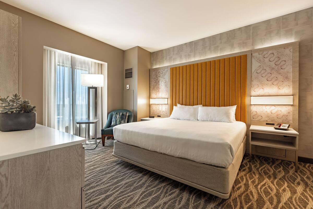 A hotel room with a large bed, a desk, a chair, a nightstand, and a potted plant.