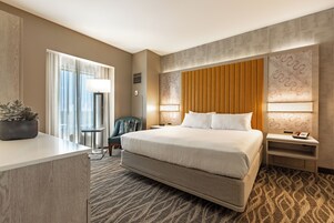 Habitación de hotel con una cama grande, un escritorio, una silla, una mesita de noche y una planta en maceta.