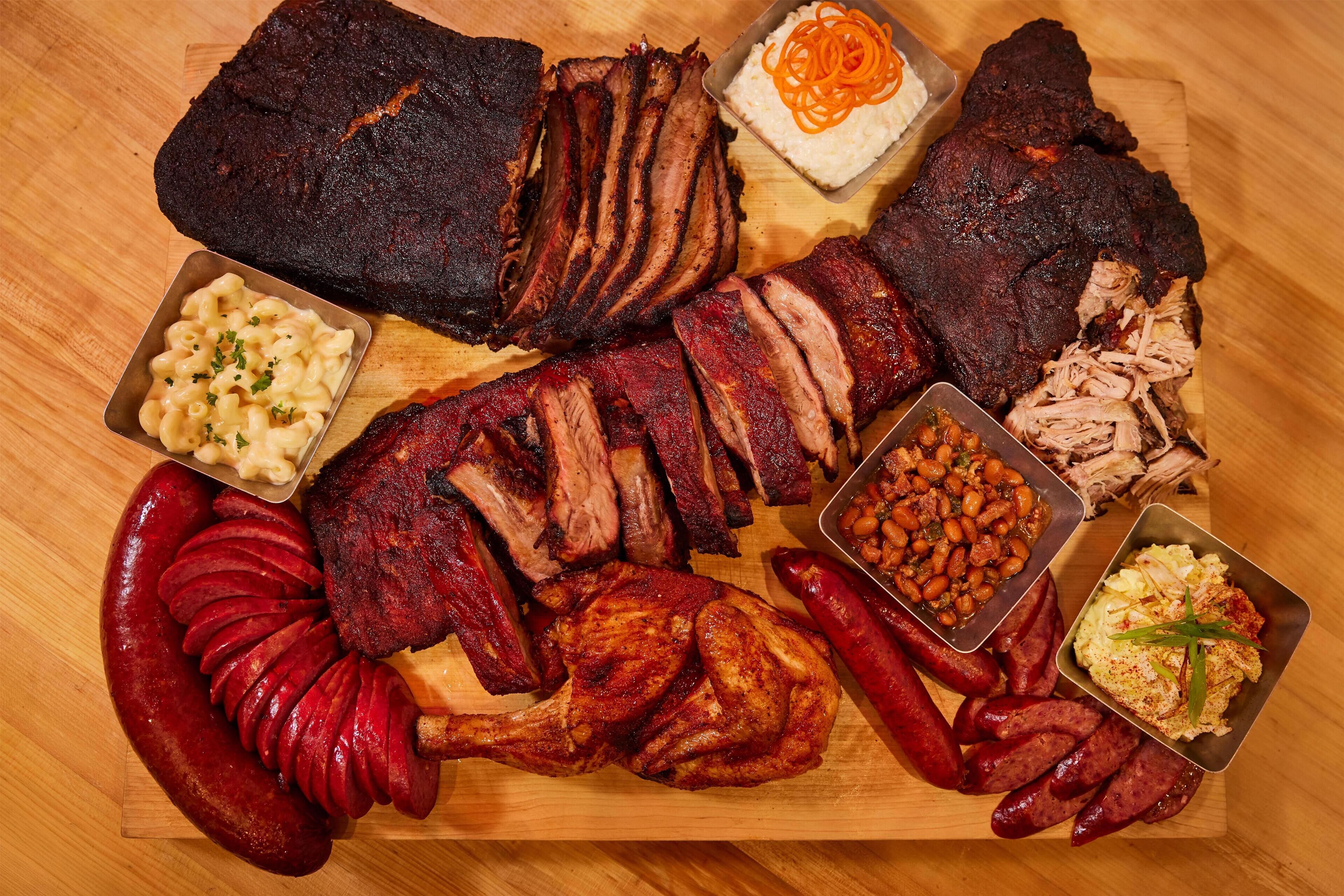 Variedad de carnes, incluyendo costillas, salchichas y un pollo entero asado, servidas sobre una tabla de madera con guarniciones.