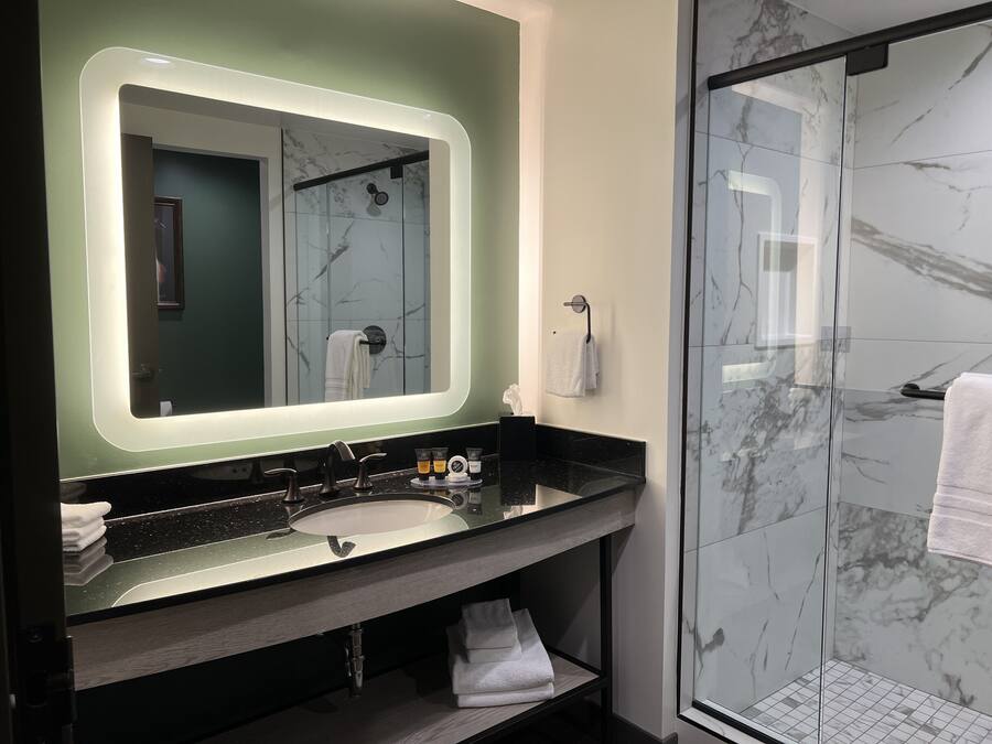 A modern bathroom with a marble shower, a large mirror with under-cabinet lighting, and a black countertop with toiletries.