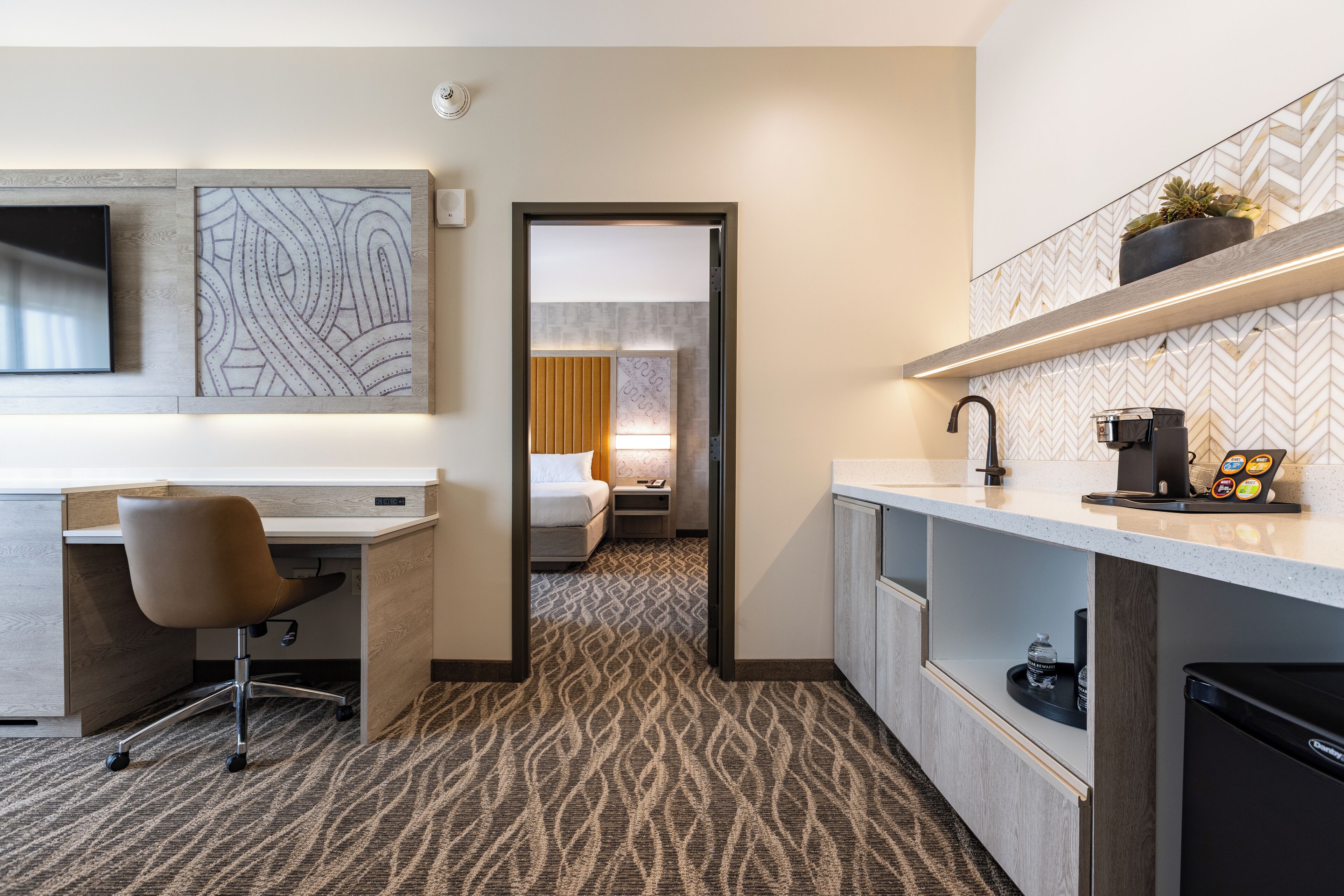 A modern hotel room with a kitchenette, a desk, a chair, a bed, and a bathroom visible through an open door.