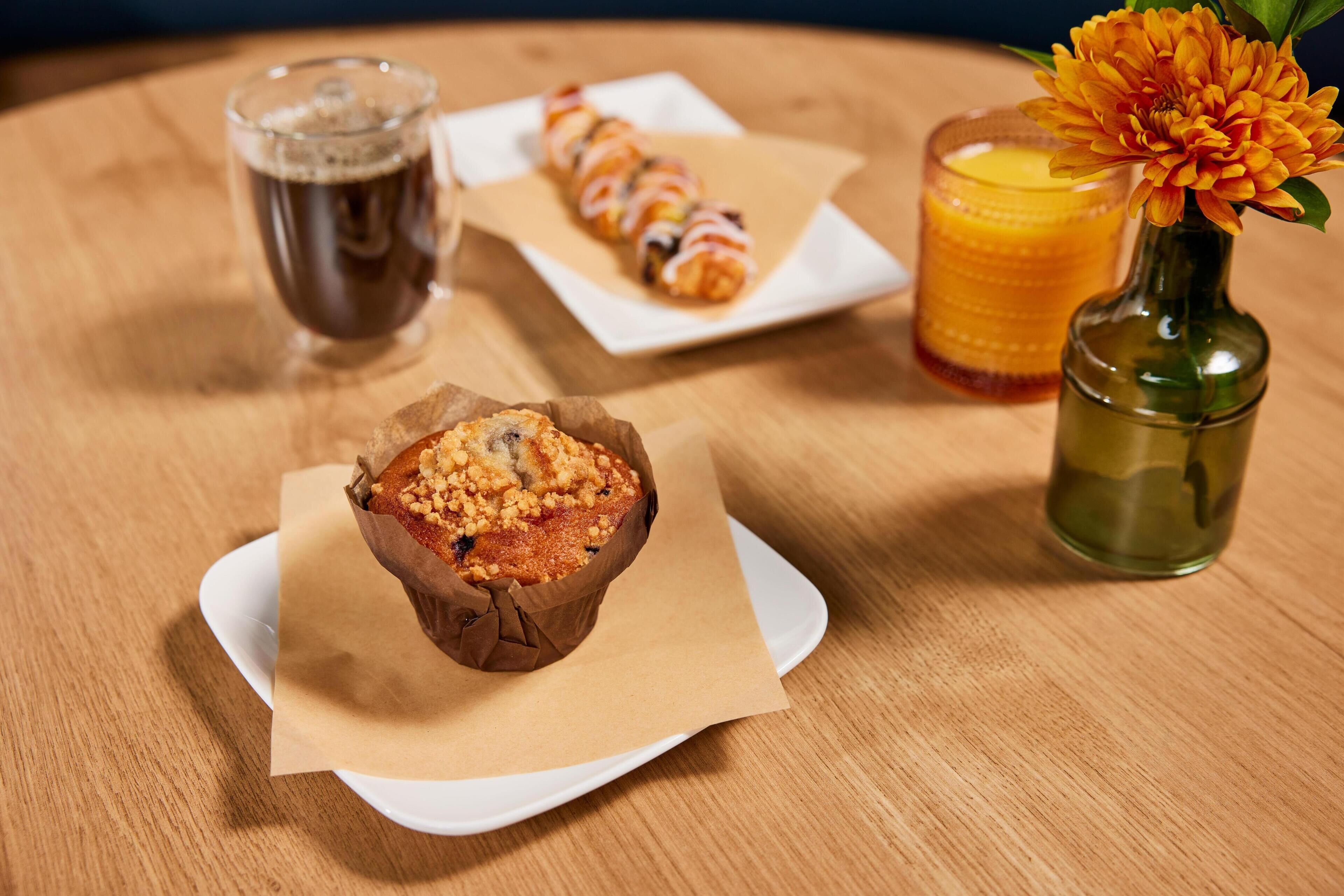 A muffin in a paper cup on a white plate, a glass of orange juice, a glass of dark beverage, and a small vase with a flower on a wooden table.