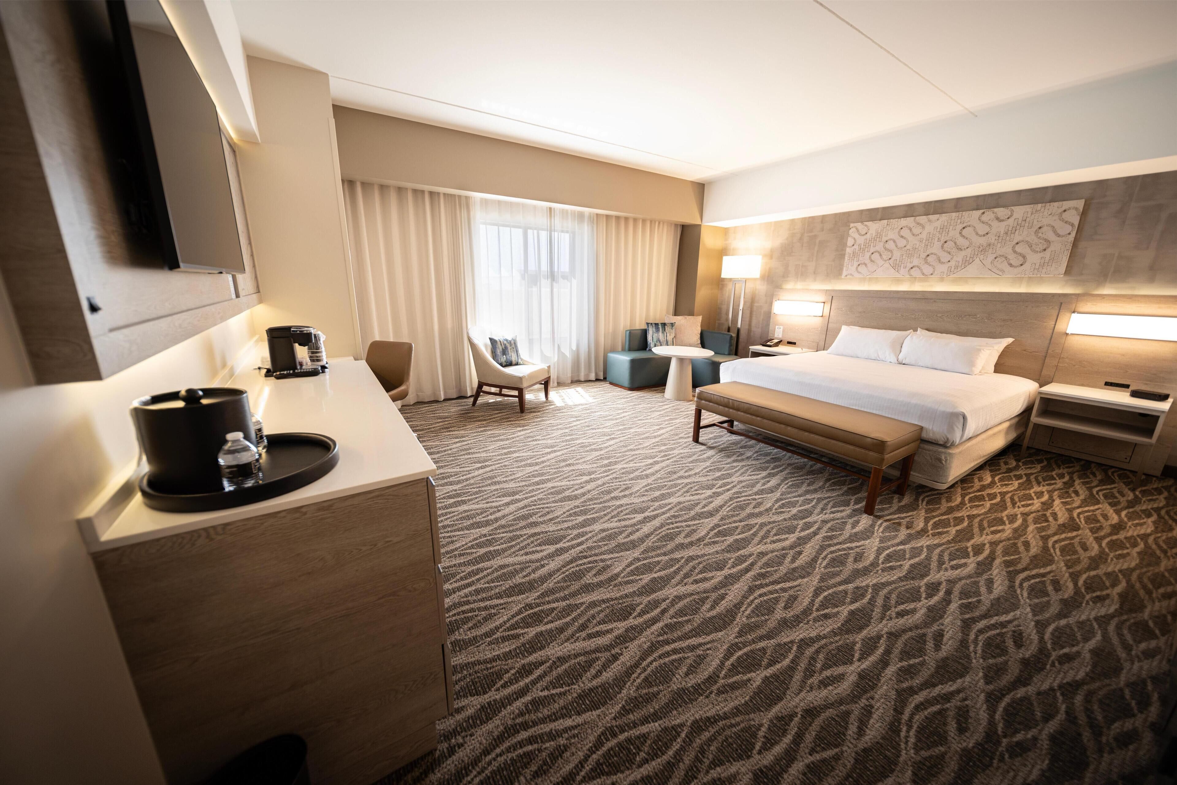 A hotel room with a bed, a desk with a coffee maker, a chair, a small table, and a TV mounted on the wall.