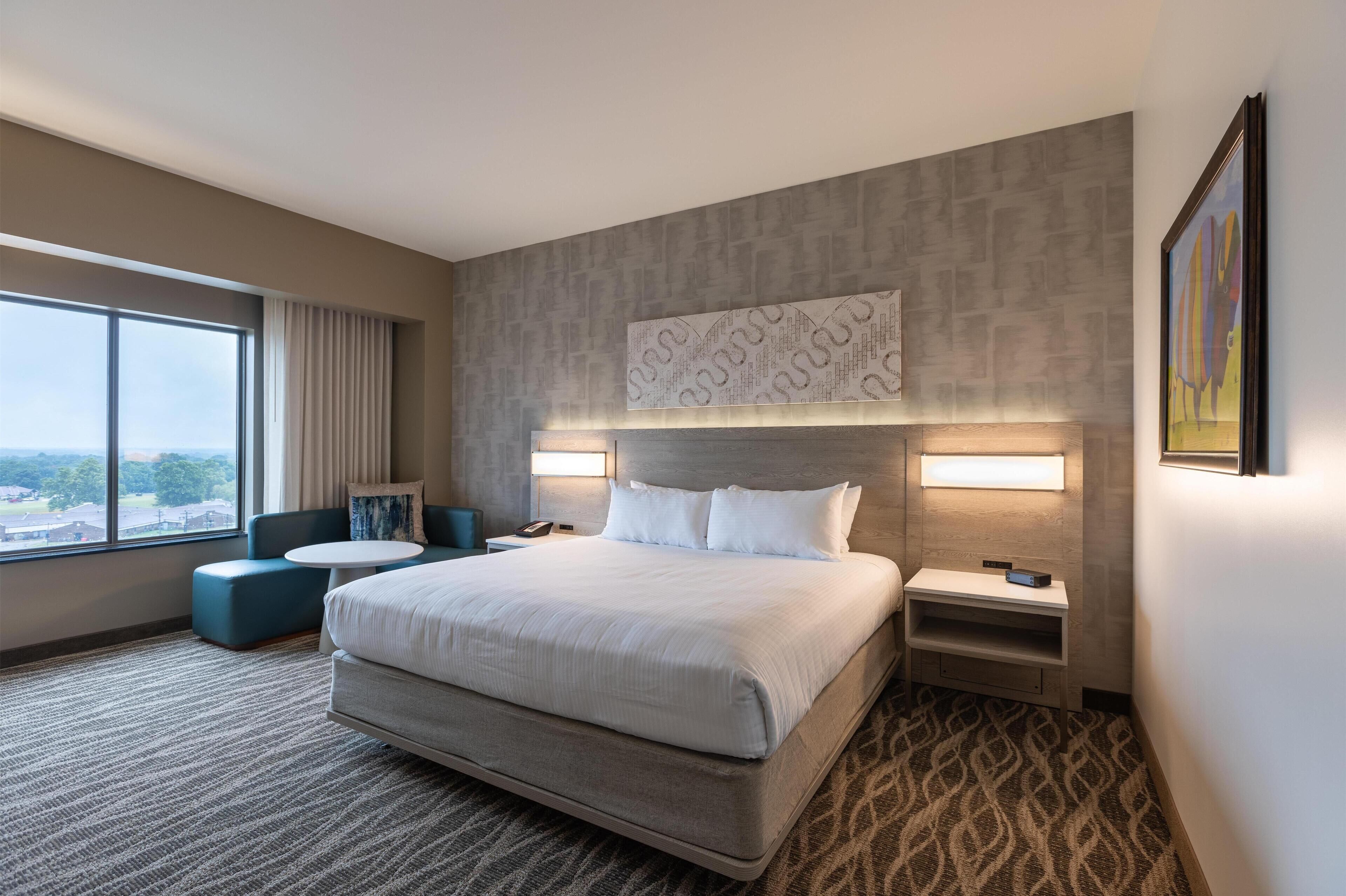 A modern hotel room with a large bed, a seating area, a desk, and a view of the city.