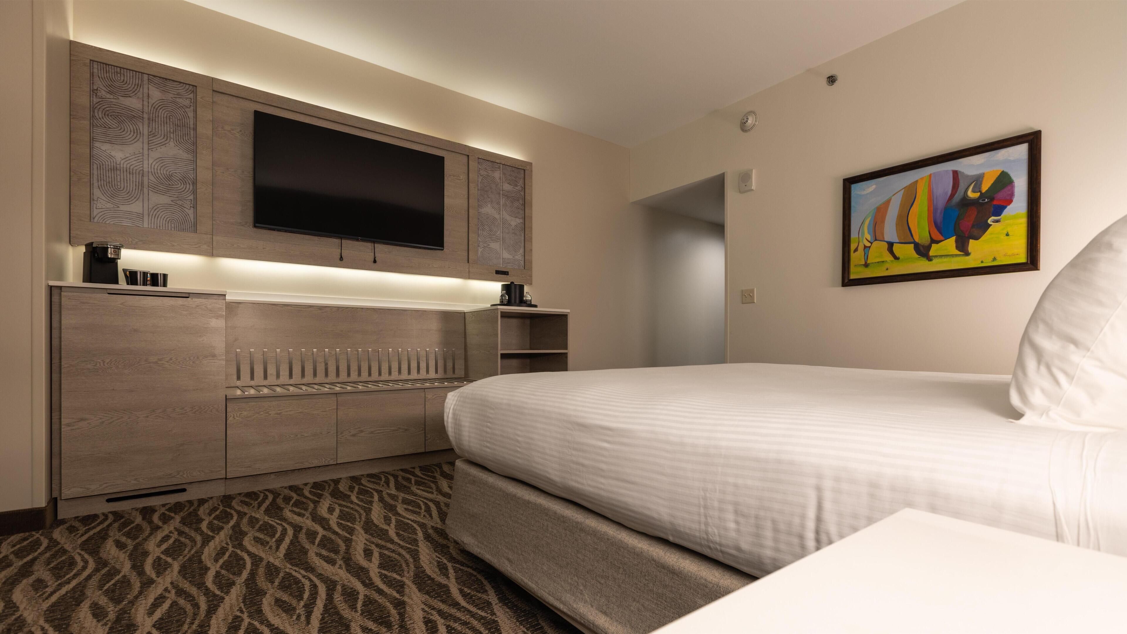 A hotel room with a bed, a flat-screen TV mounted above a wooden cabinet, a painting on the wall, and a patterned carpet.