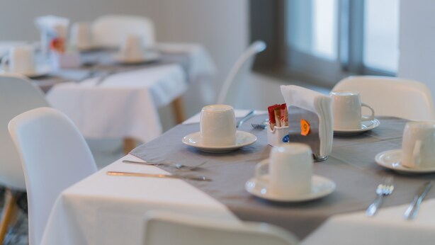 Un comedor con mesas dispuestas para el desayuno, incluyendo platos, tazas y cubiertos.