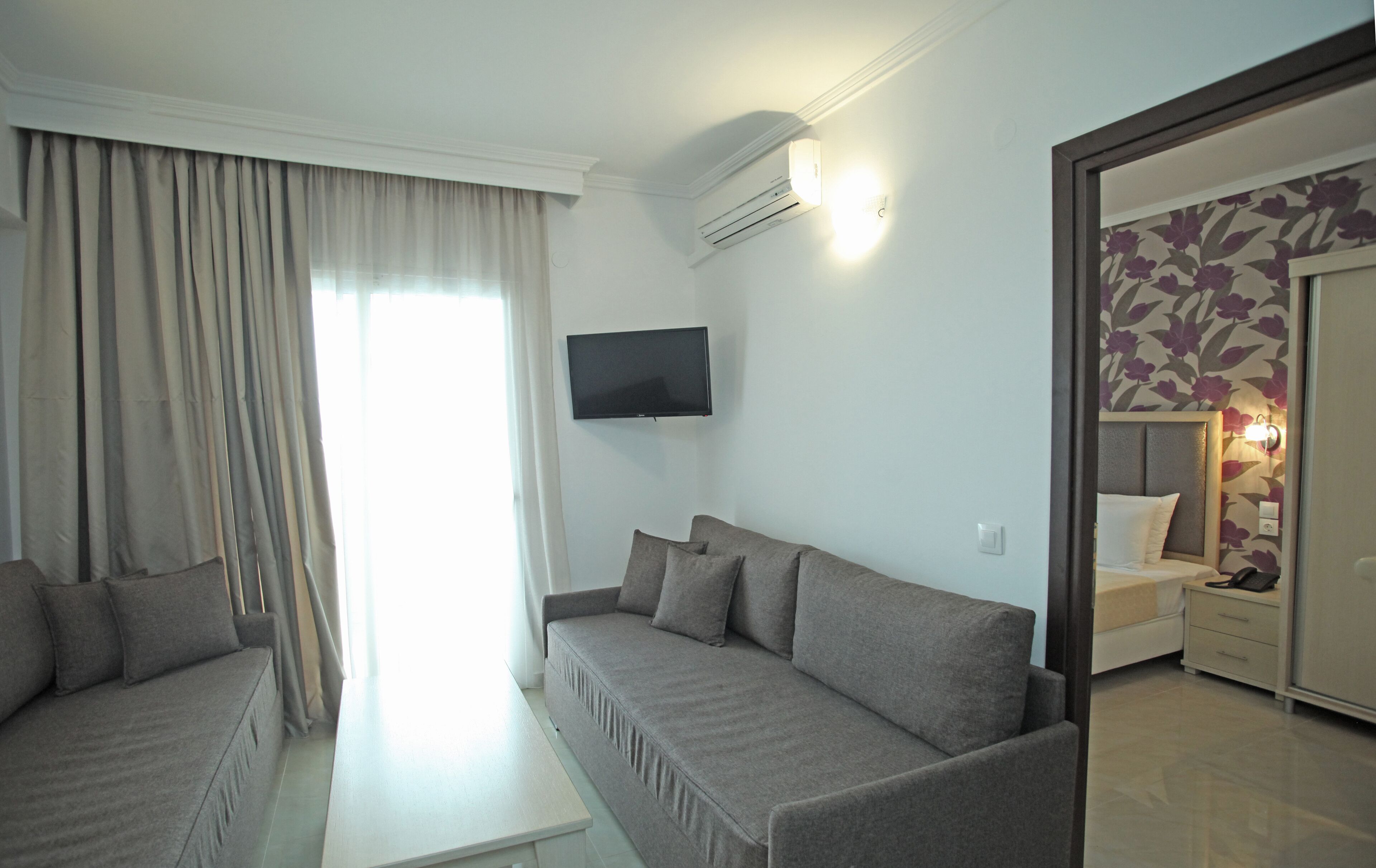 Una habitación de hotel moderna con un sofá gris, un televisor de pantalla plana montado en la pared y una cama con cabecera estampada con motivos florales.