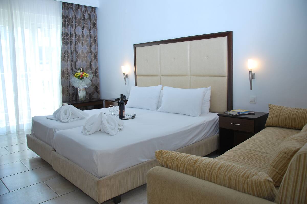 A hotel room with a large bed, a bedside table with a lamp, a sofa, and a window with curtains.