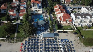 Vista aérea de un resort con una gran piscina, varias edificaciones y una zona de playa con numerosos paraguas.