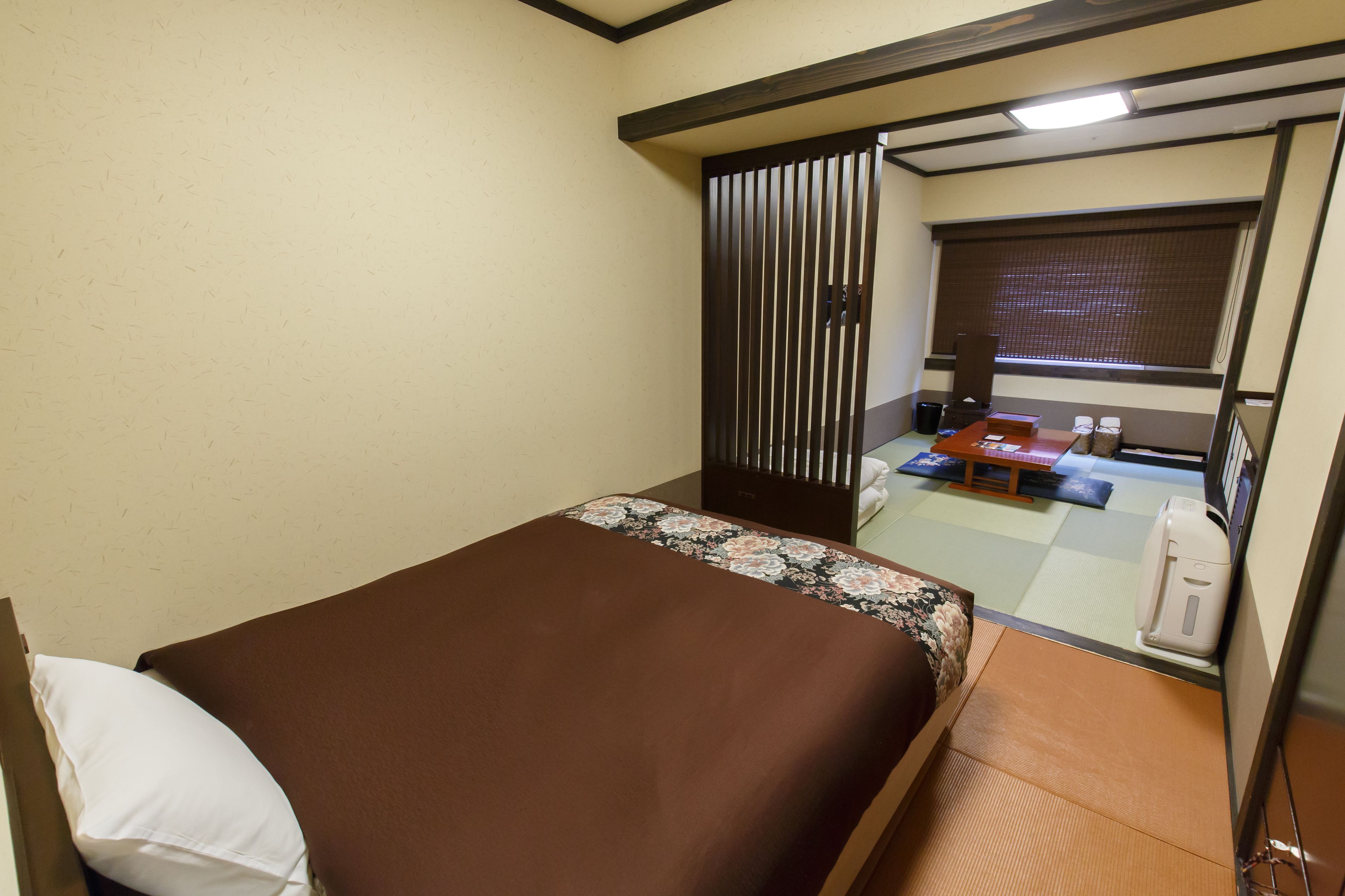 Een hotelkamer met een bed, een klein tafeltje, een waterkoker en een raam met jaloezieën.
