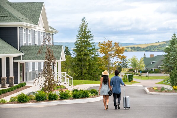 A couple walking on a residential street with a house featuring a green roof, white trim, and a view of a lake.
