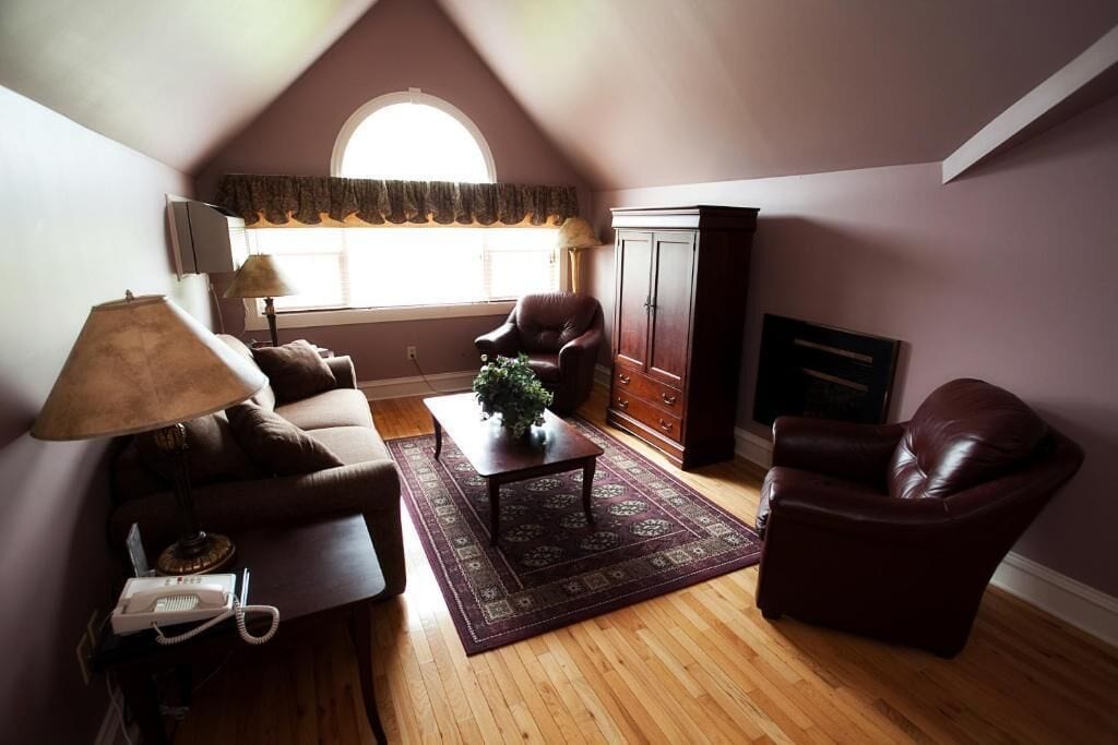 A cozy living room with a brown leather sofa, a coffee table with a plant, a fireplace, and a large window with a decorative valance.