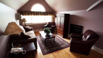A cozy living room with a brown leather sofa, a coffee table with a plant, a fireplace, and a large window with a decorative valance.