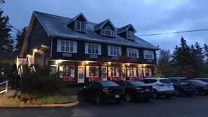 Un bâtiment à deux étages, doté d’une façade sombre et de nombreuses fenêtres, avec une véranda comprenant des sièges extérieurs et un parking où sont stationnées plusieurs voitures.