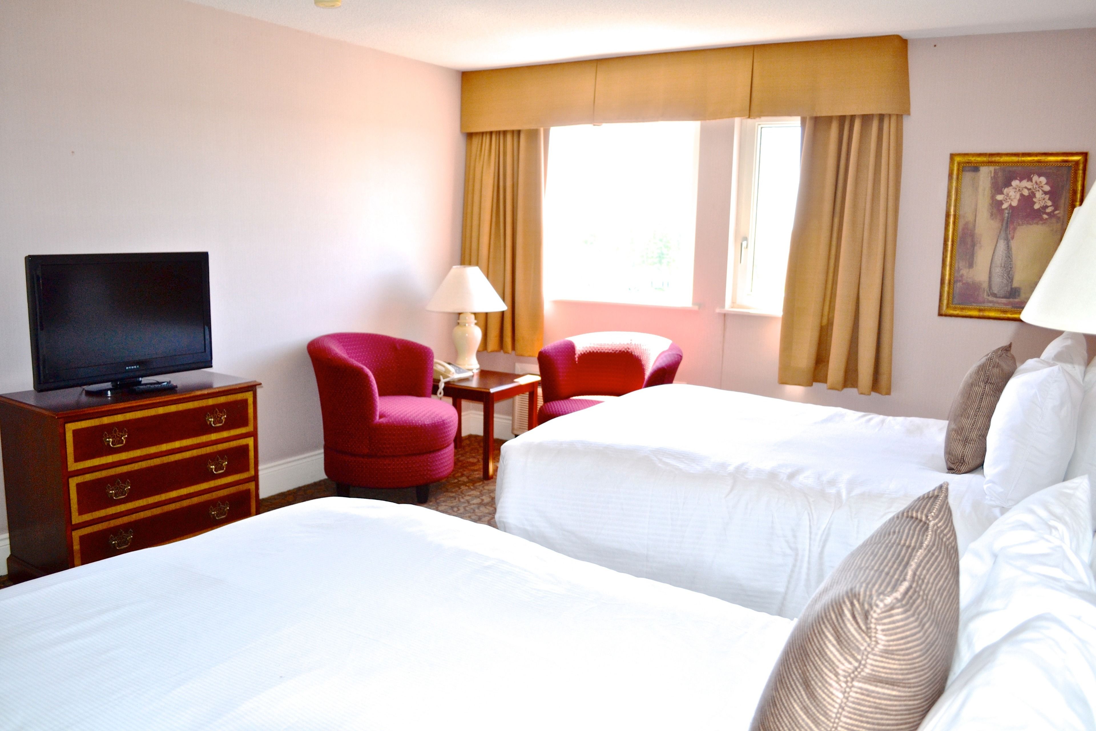 A hotel room with two beds, a television on a wooden dresser, a red armchair, a small table with a lamp, and a window with curtains.