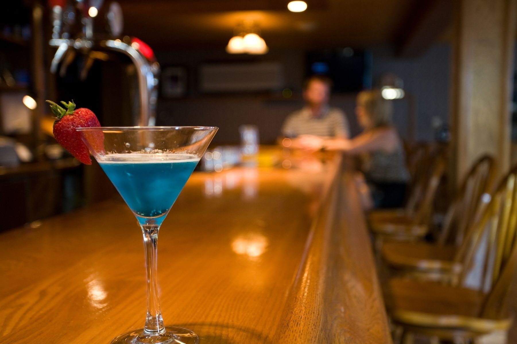 Un verre à martini contenant un liquide bleu et décoré d’une fraise, posé sur le comptoir en bois d’un bar.