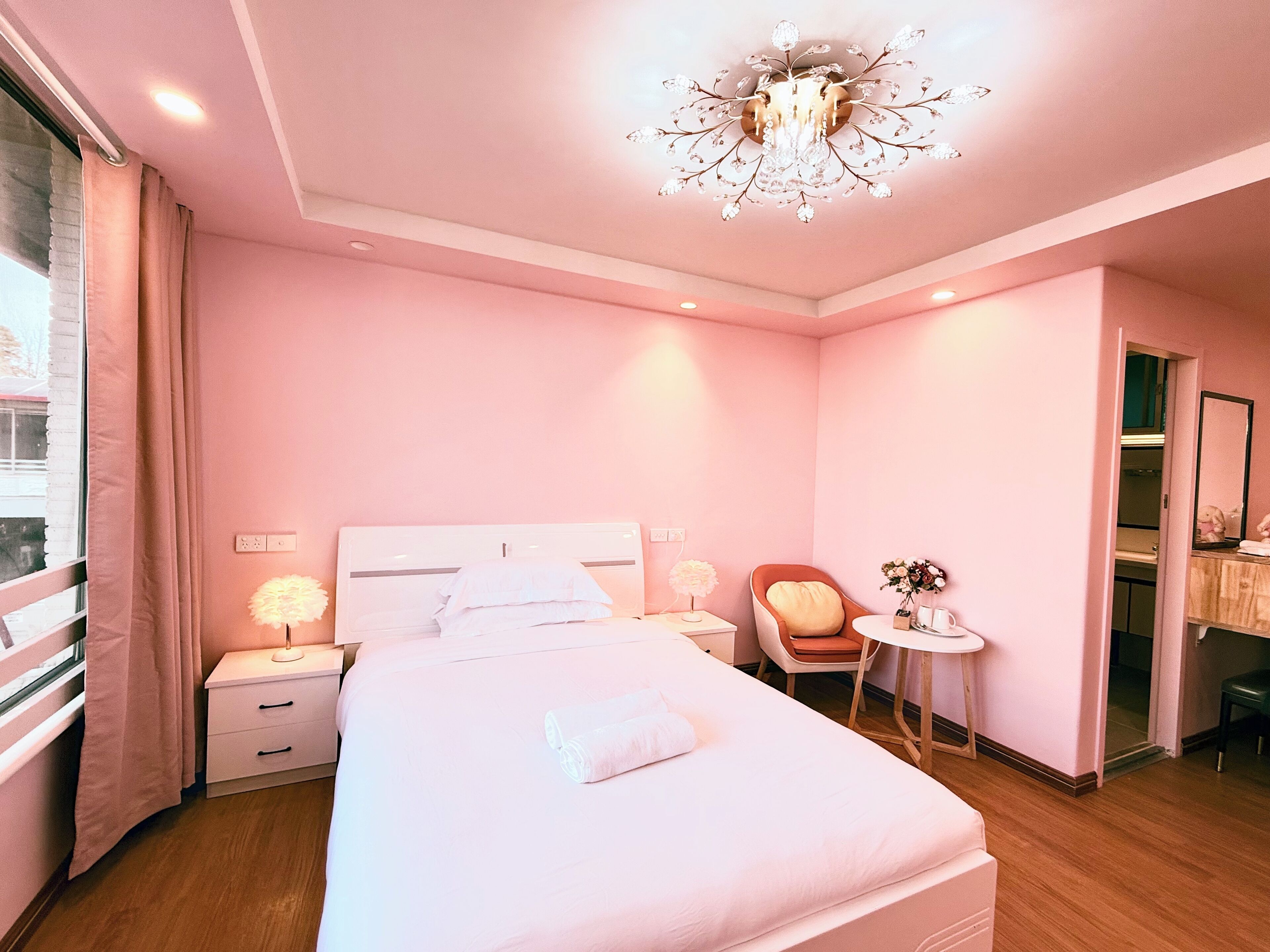 Habitación de hotel en tonos rosados, con una cama grande, lámparas de noche, una mesita con un florero y una silla.