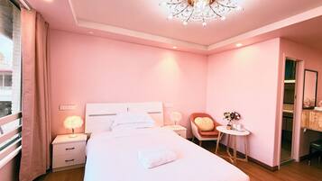 Habitación de hotel en tonos rosados, con una cama grande, lámparas de noche, una mesita con un florero y una silla.