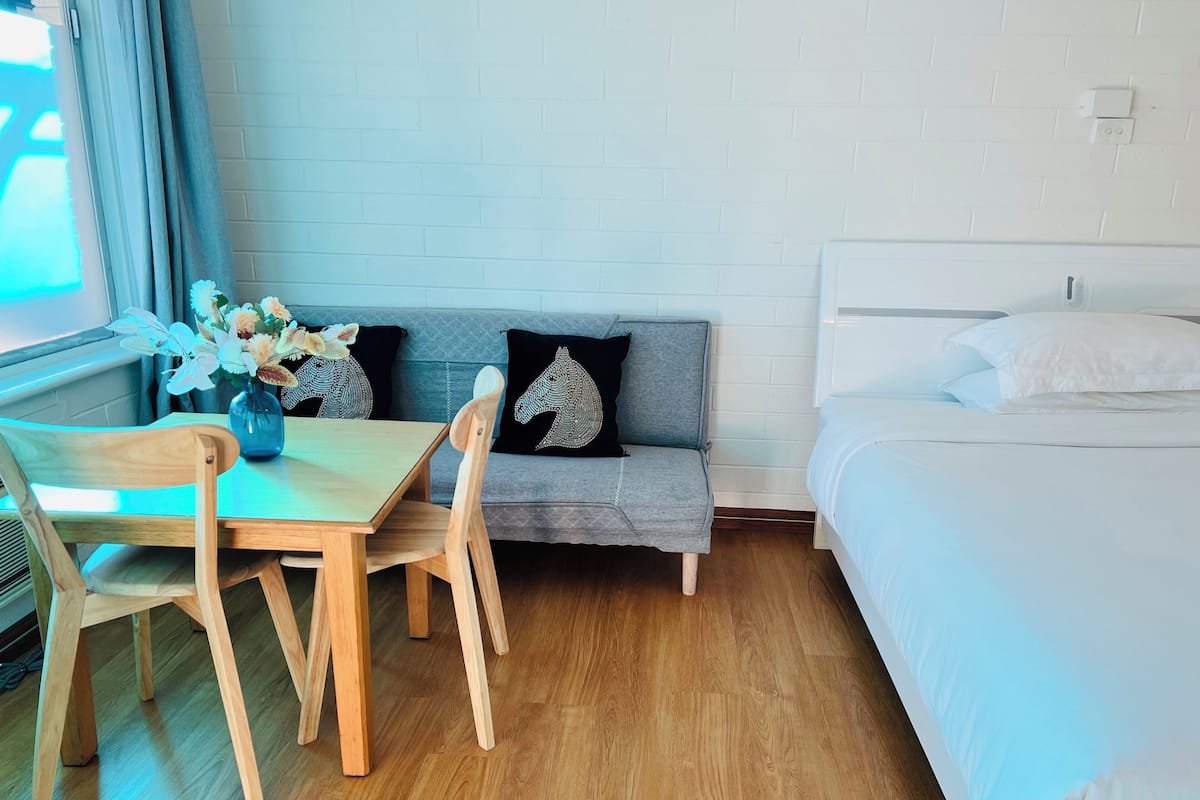 A room with a wooden table, two chairs, a sofa with decorative pillows, a bed, and a window with curtains.