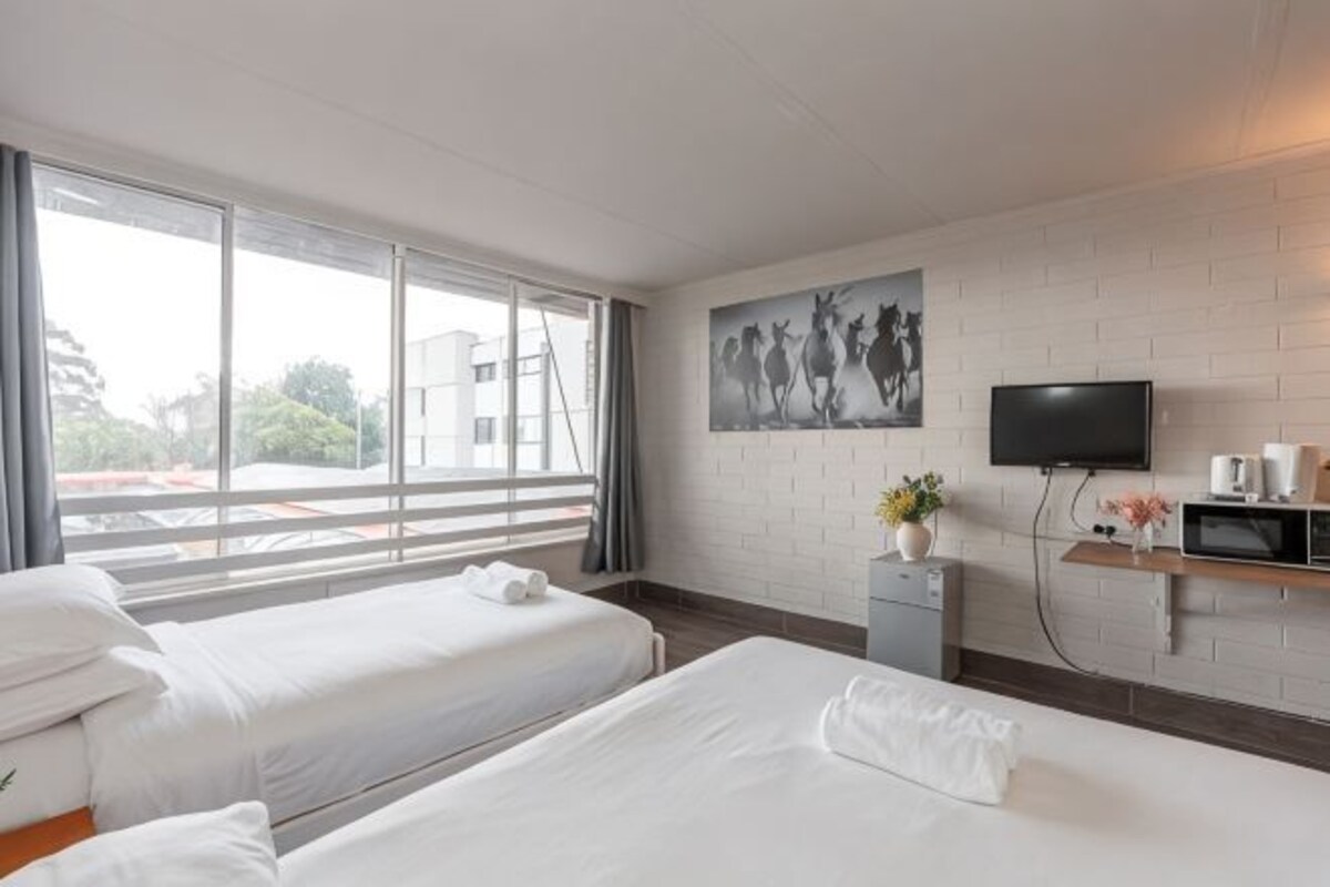 A hotel room with two beds, a TV, a small desk, and a window with a view of buildings.