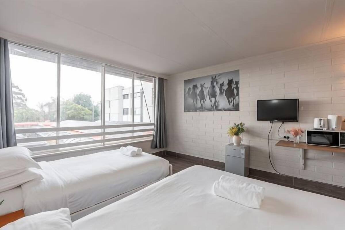 A hotel room with two beds, a TV, a small desk, and a window with a view of buildings.