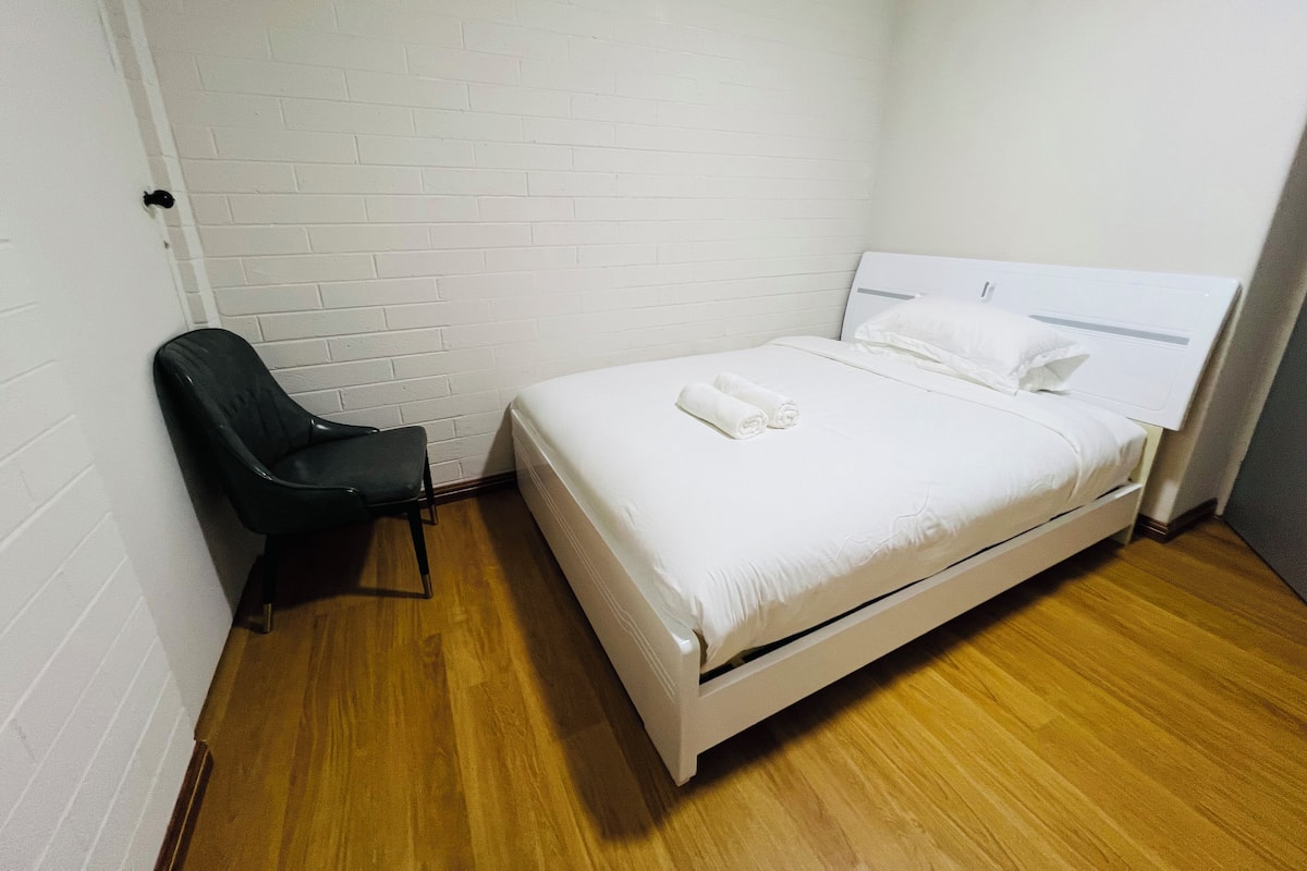 A small, clean room with a single bed, a chair, and white brick walls.