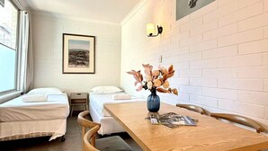 Habitación de hotel con dos camas individuales, una mesa de madera con un jarrón de flores, un cuadro en la pared y una ventana con cortinas.