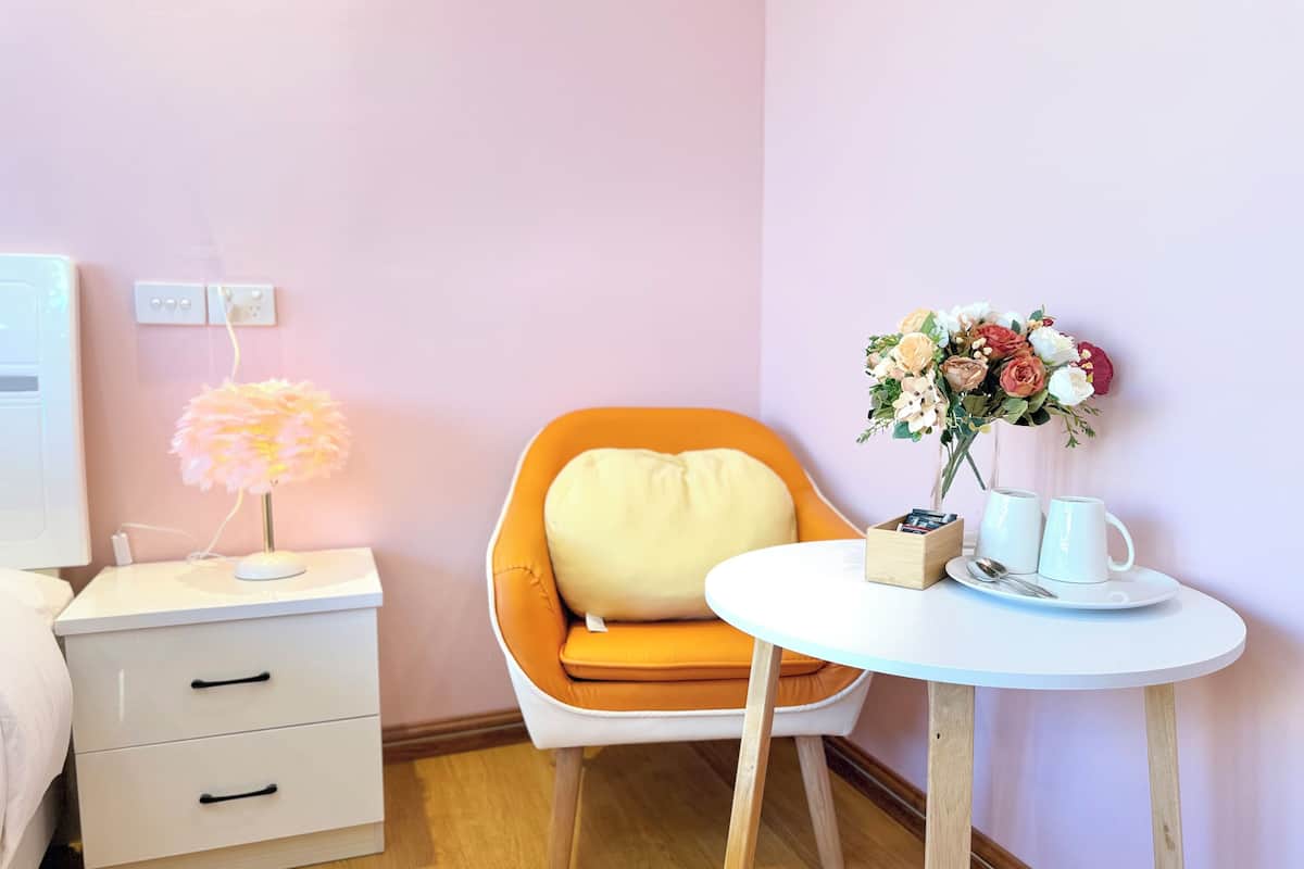 A neatly arranged hotel room with a bed, a bedside table with a lamp, a chair, a small table with a vase of flowers, and a wall socket.